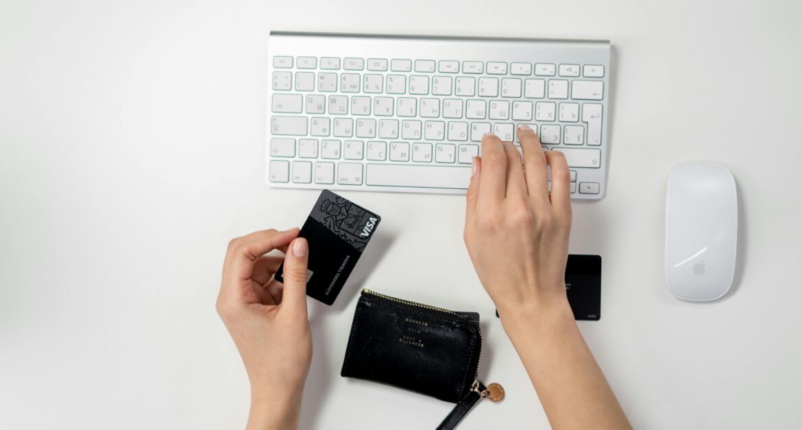Hands typing on an Apple keyboard with a credit card in hand, next to a wallet and a mouse on a white surface.