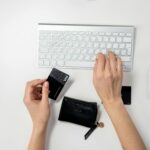 Hands typing on an Apple keyboard with a credit card in hand, next to a wallet and a mouse on a white surface.